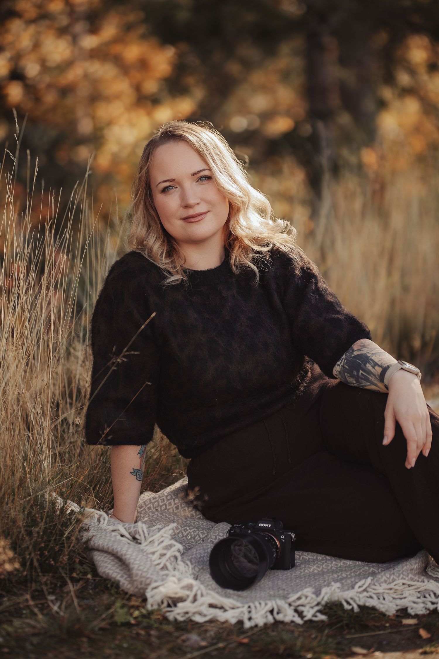 Blonde woman in black outfit sits on blanket outdoors with camera, autumn foliage background.