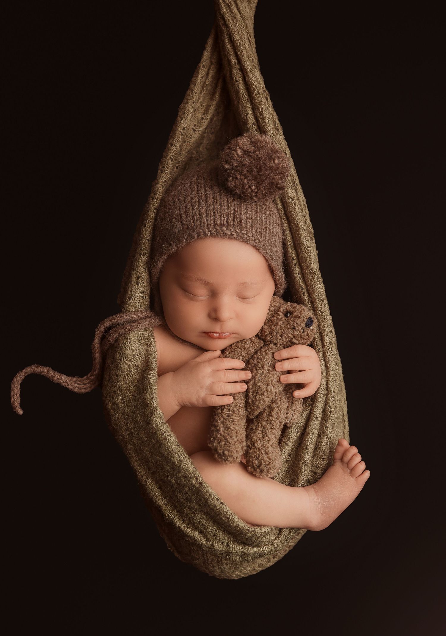 Sleeping newborn baby in green wrap cocoon wearing knit bonnet, cradling a brown teddy bear against dark background.