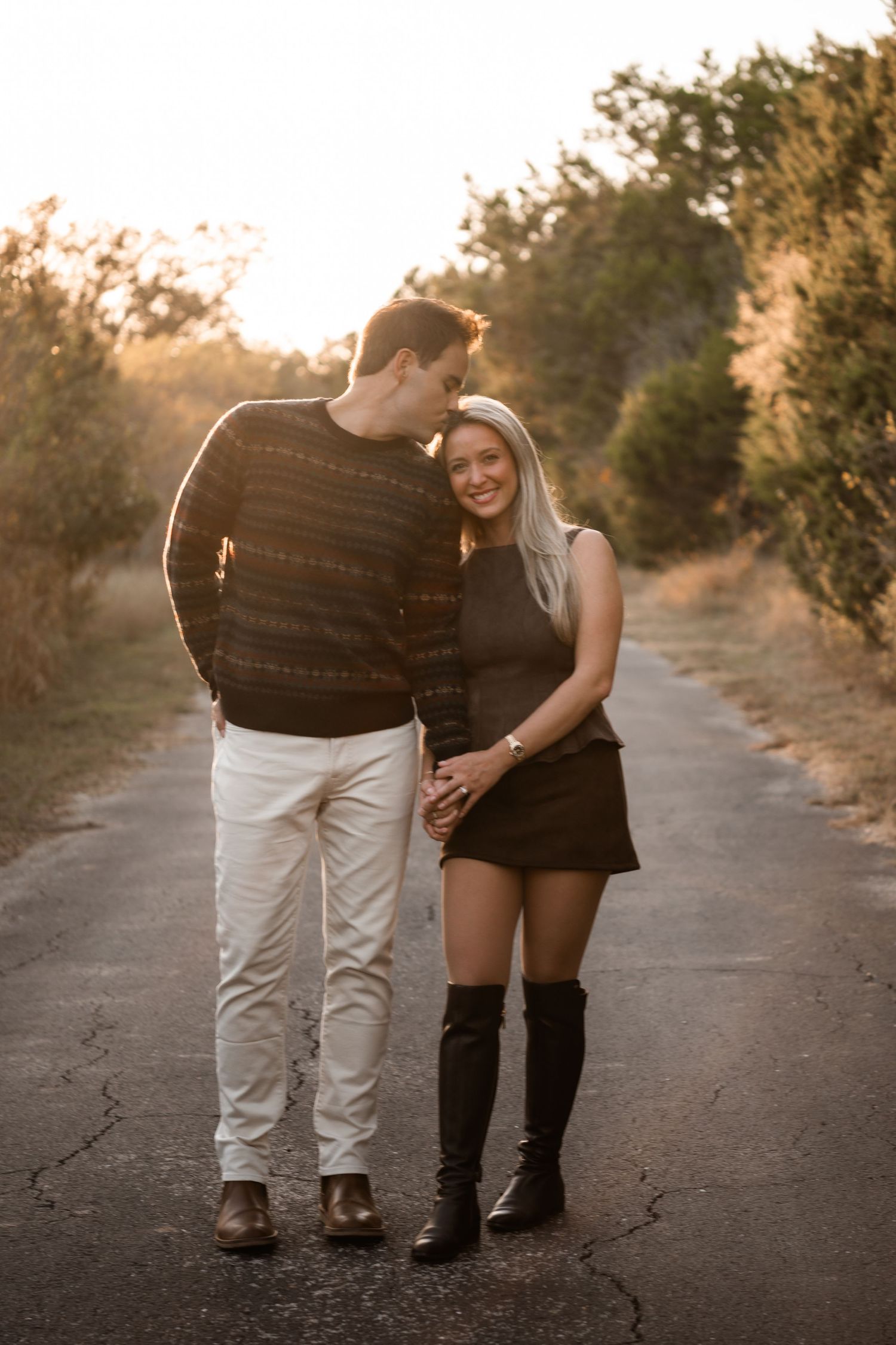 Couple holding hands on a sunlit road at golden hour, woman in black mini skirt and knee-high boots, man in patterned sweater.