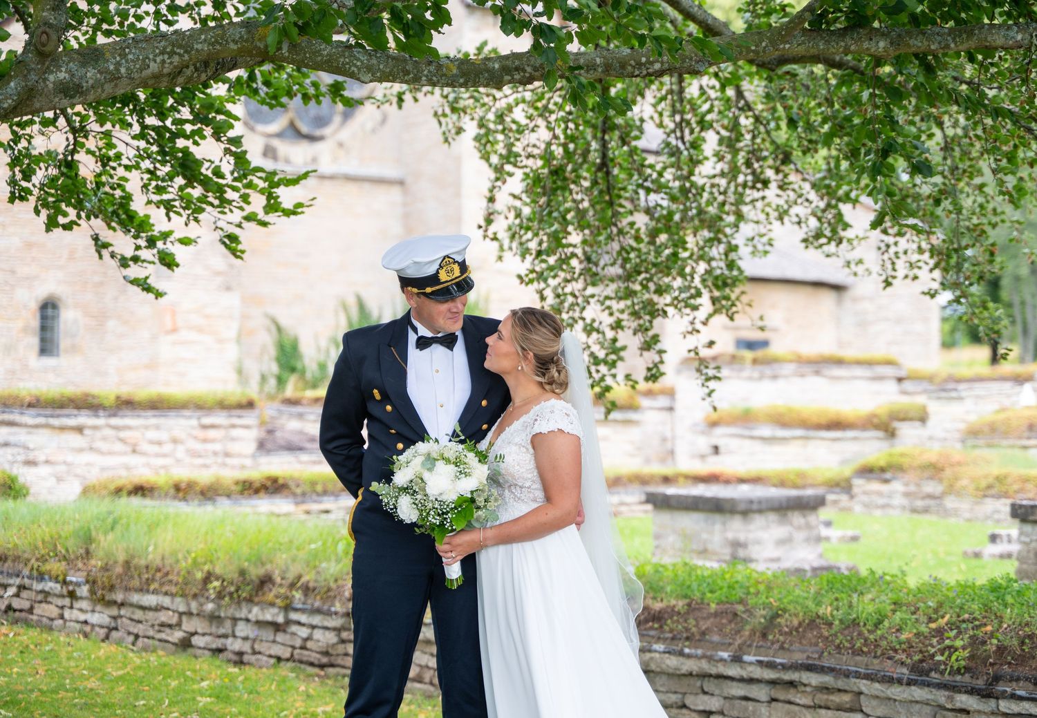 A bride and groom share a romantic moment outdoors, with the groom in a naval uniform and the bride holding a white bouquet.