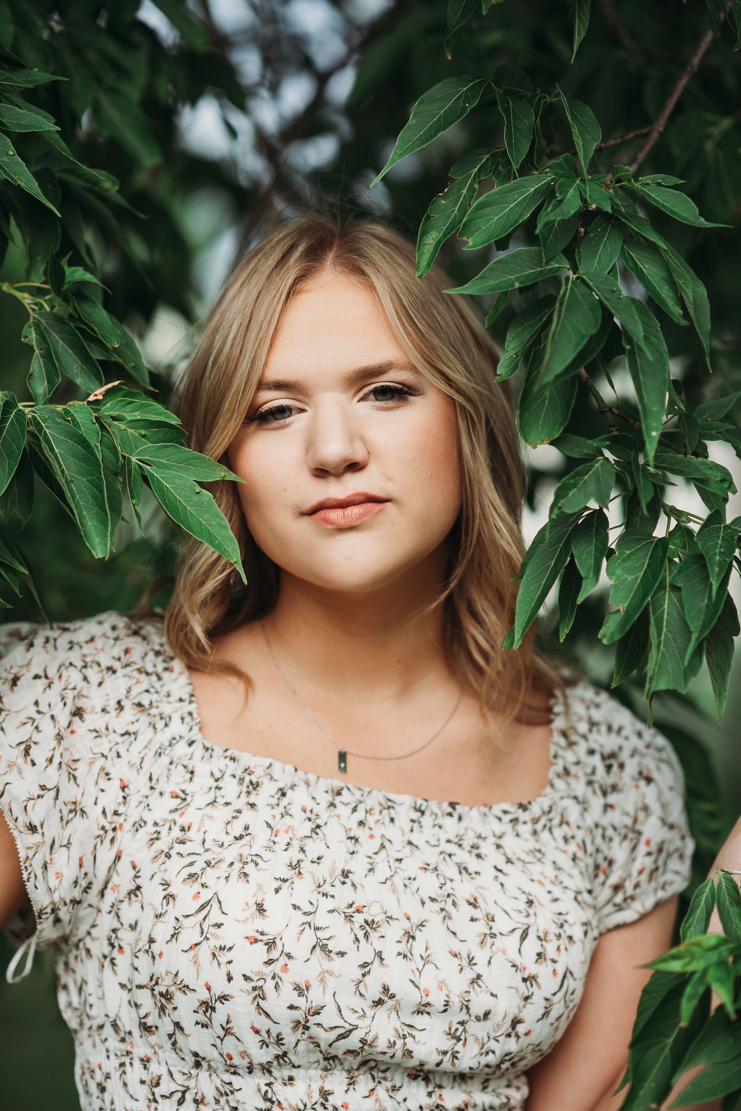 Portrait framed by green leaves featuring floral top in natural outdoor setting.
