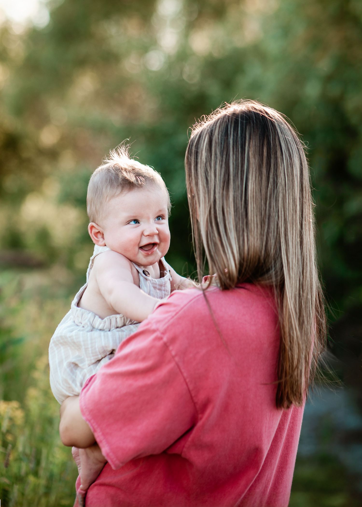 Baby in white outfit smiles while being held outdoors in natural lighting.