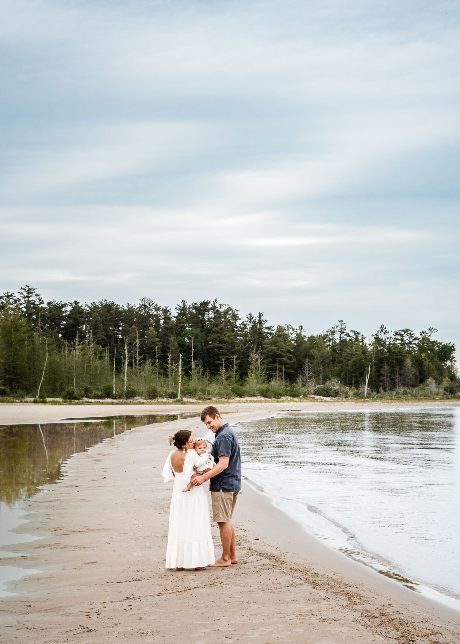 A couple shares an intimate moment on a serene beach with cloudy skies and trees lining the shore.