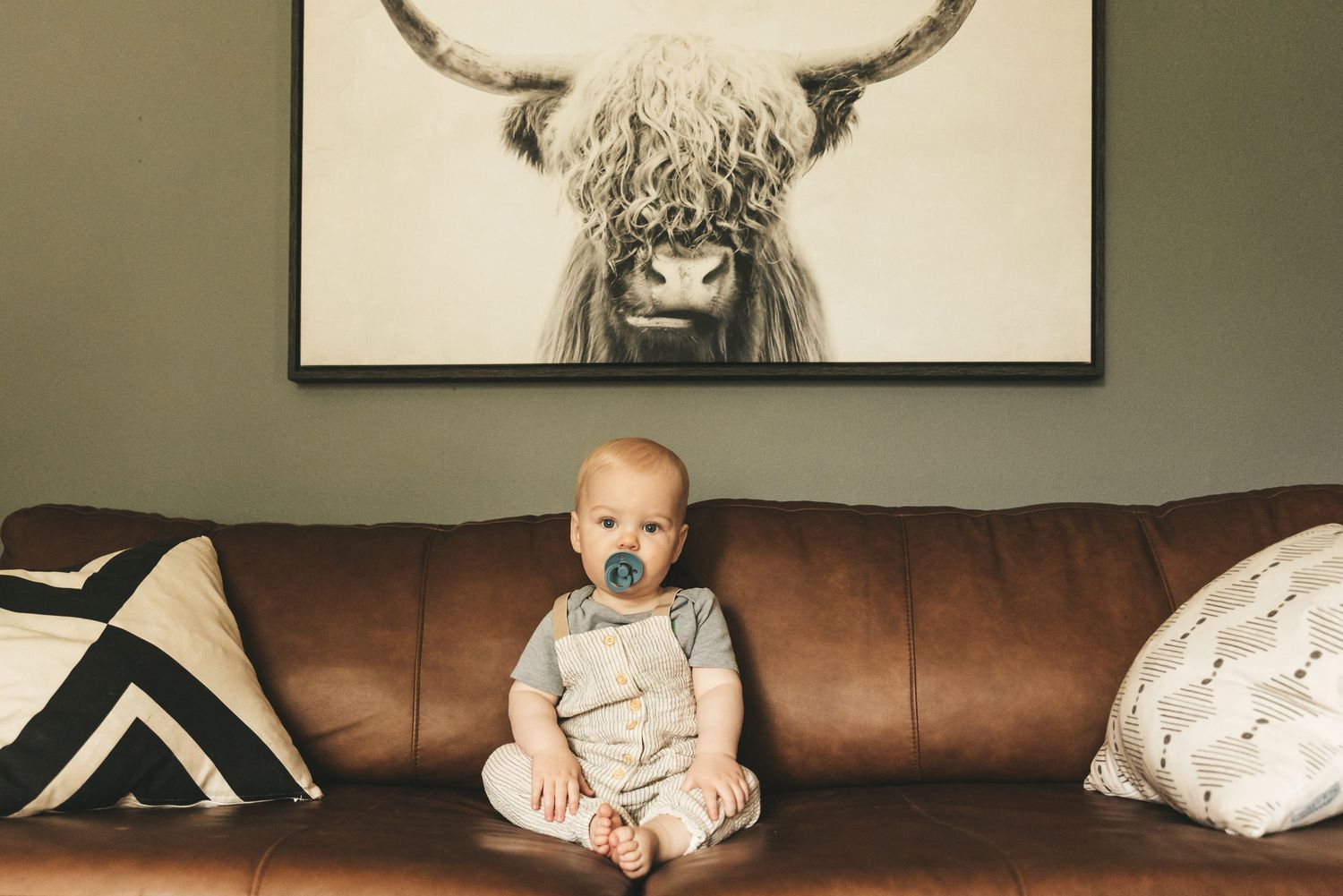 Small child sitting on brown leather couch beneath framed highland cow artwork.