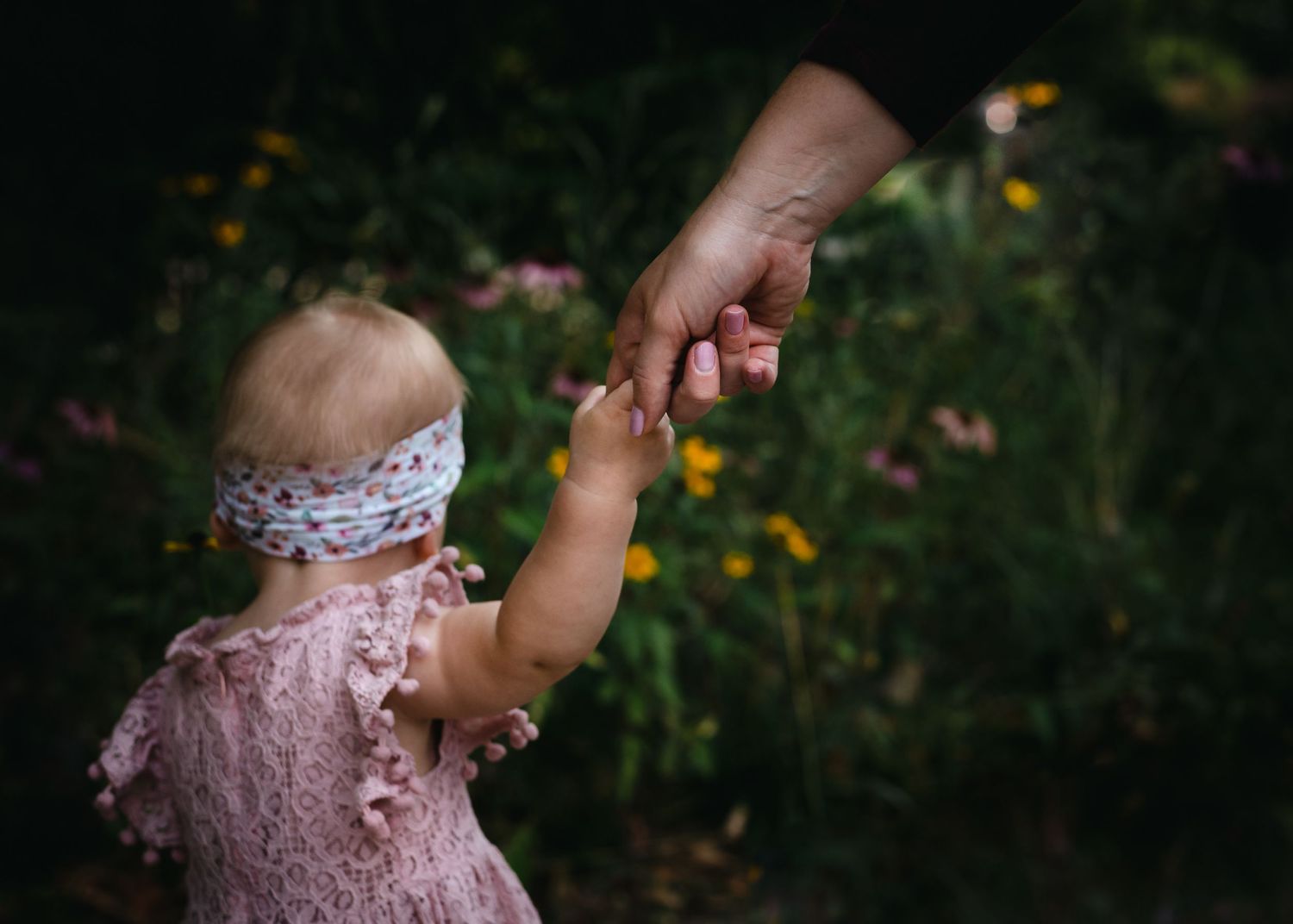 A small child in a pink dress and headband holds hands with someone in a garden setting during sunset.