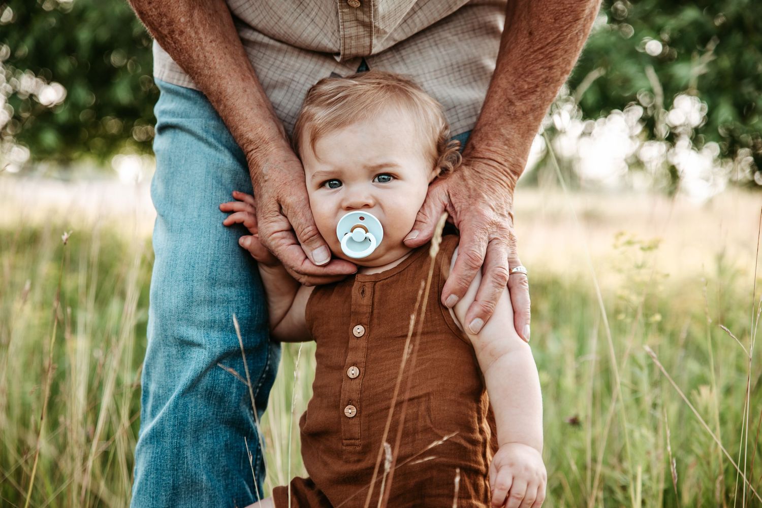 Close-up of hands holding small child in brown overalls in grassy field.