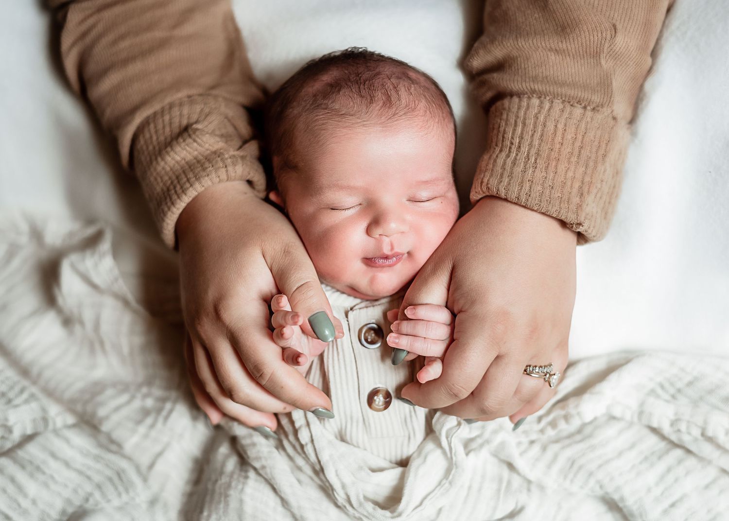 Gentle hands cradle a sleeping newborn wearing white against a soft white blanket.