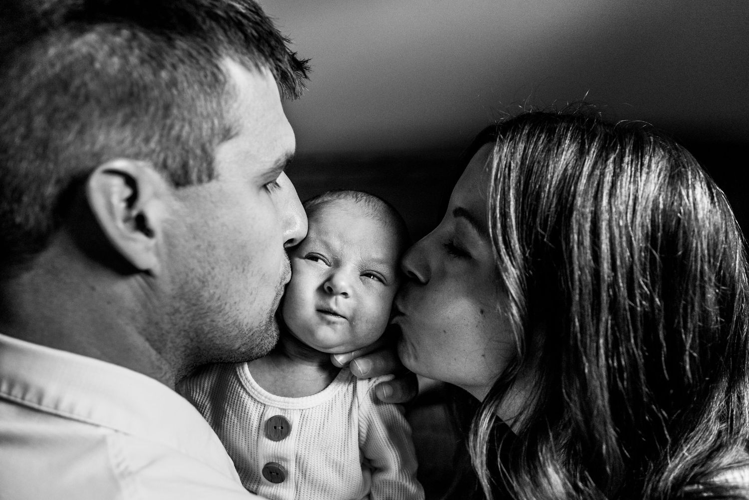 Lifestyle black and white family moment with baby being kissed by parents.