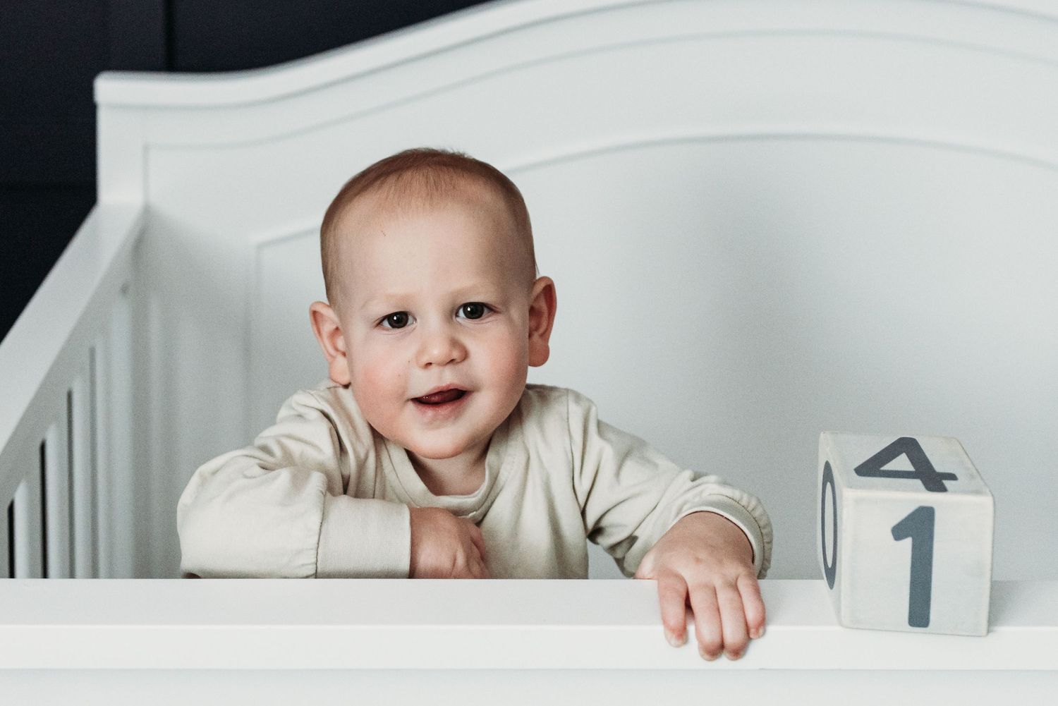 Smiling baby in white onesie leans on crib rail next to number one block.