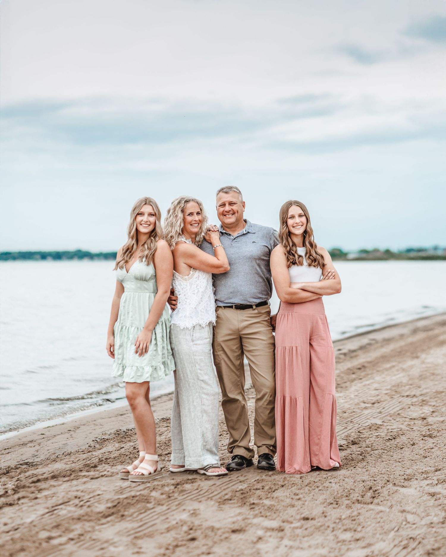 Family portrait on a beach with soft pastel dresses against cloudy sky.