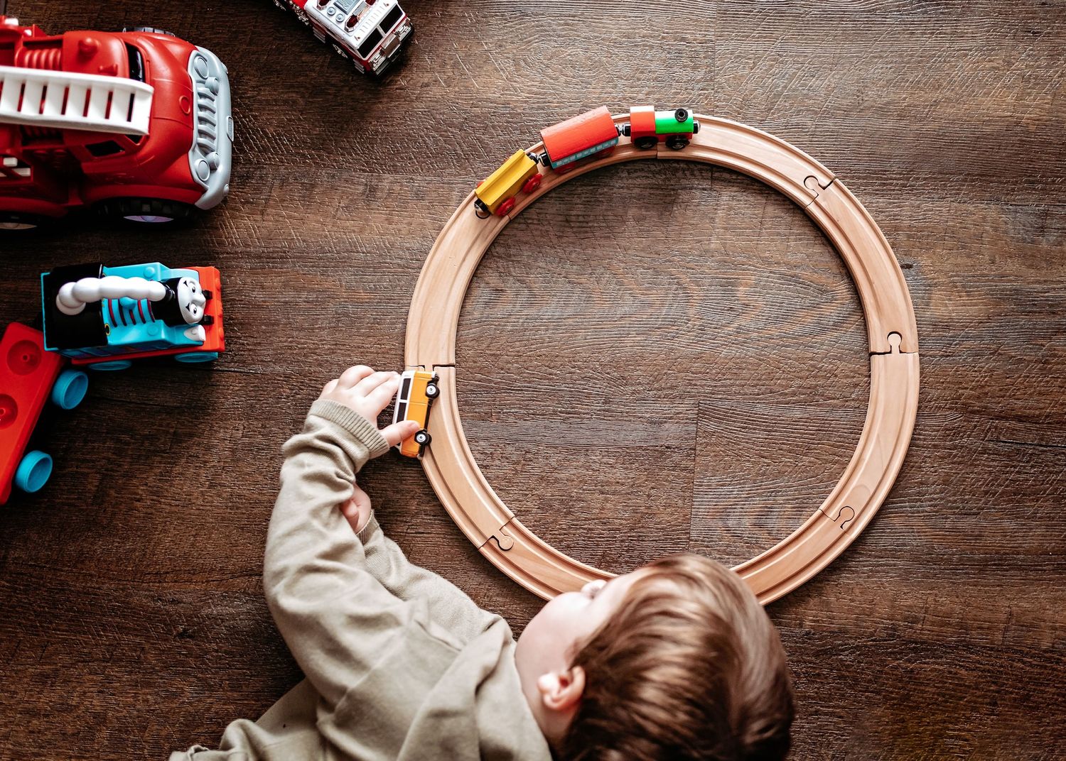 Overhead view of child playing with wooden train track and toys on hardwood floor.