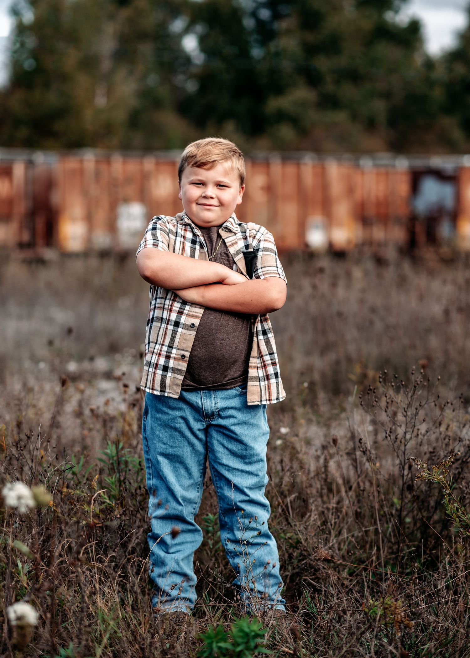 Young person in plaid shirt and jeans stands confidently in overgrown field with fence in background.