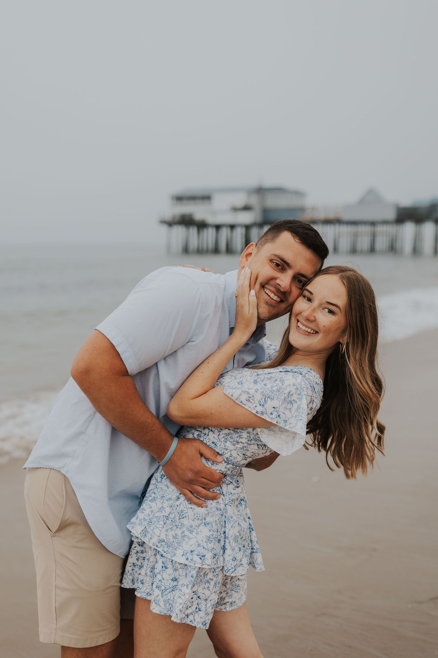 Happy couple embracing and laughing on beach with pier in background during sunset photoshoot.