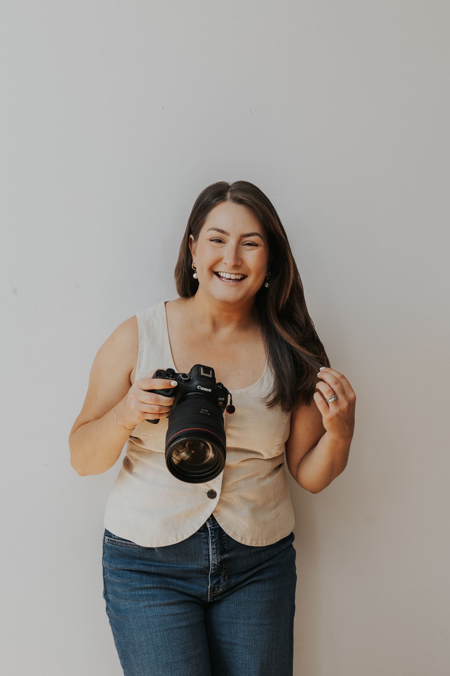 A person in jeans and a beige tank top poses casually against a white wall while holding a professional camera.