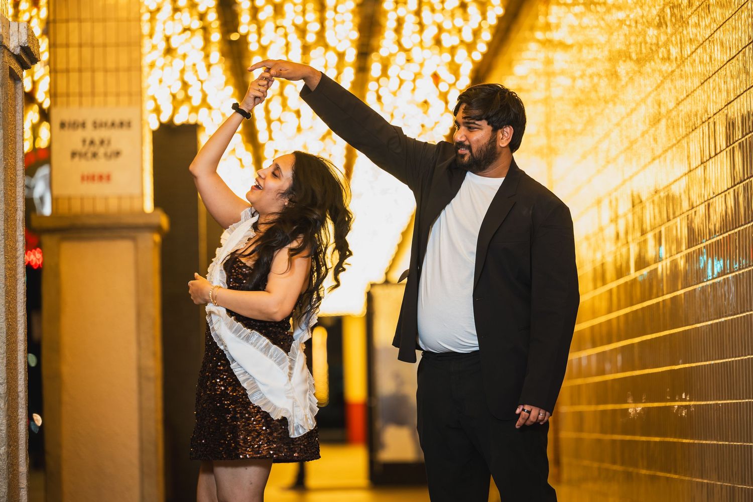 Couple dancing and spinning joyfully under golden string lights in a warmly lit corridor.