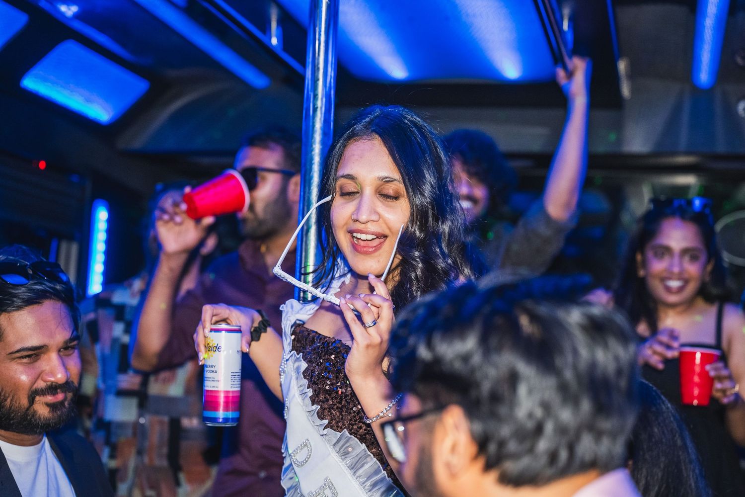 Woman wearing sash smiling and dancing on a party bus with friends holding drinks.
