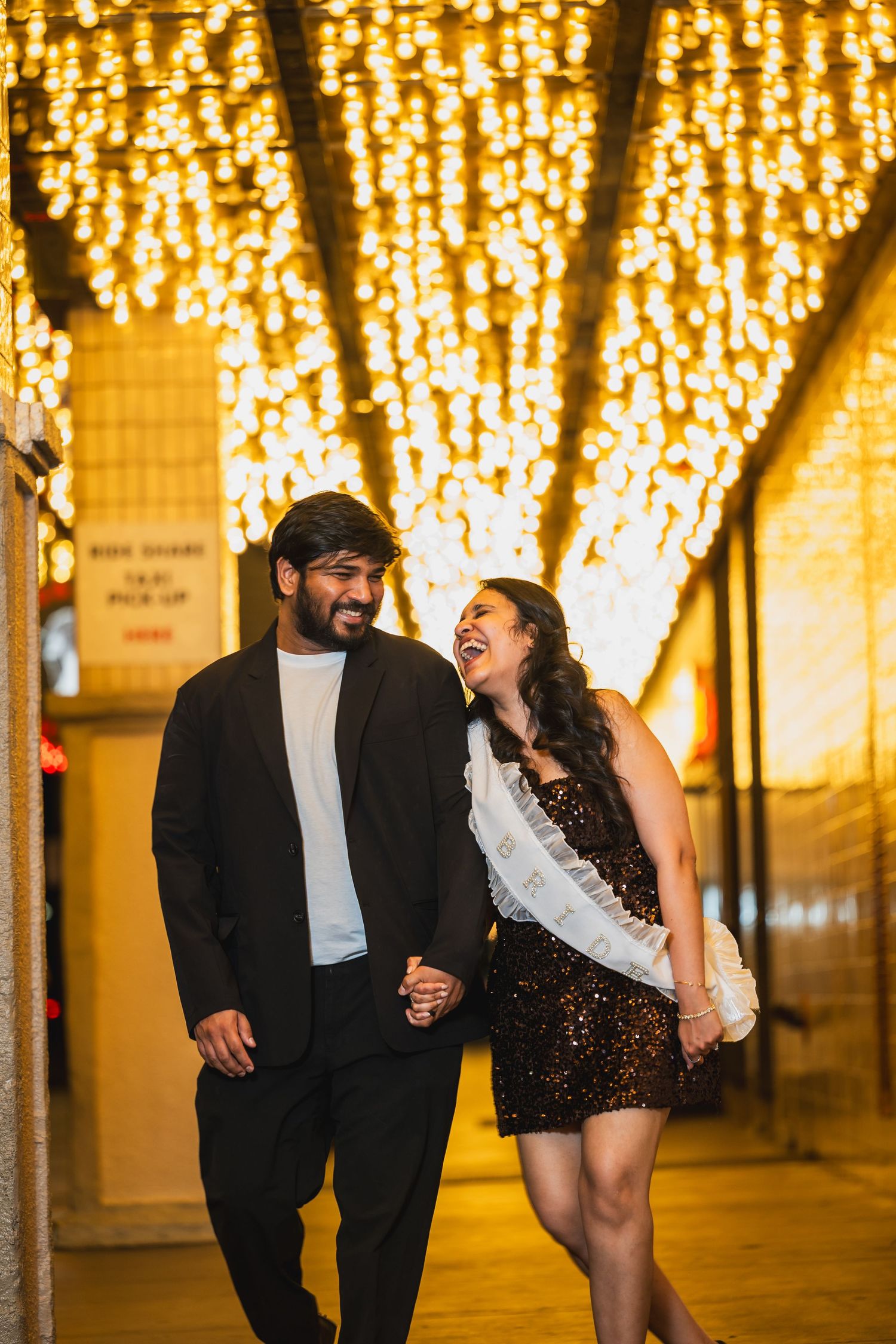 Laughing couple walking hand in hand under warm cascading string lights at night.