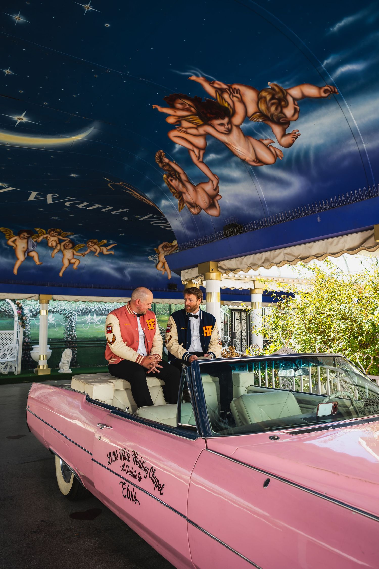 Two men seated in a classic pink convertible beneath an ornate painted mural at Elvis chapel.