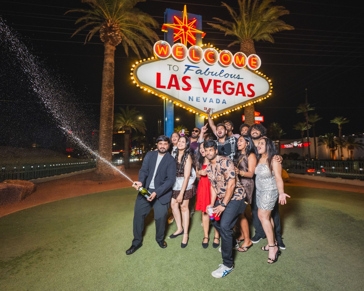 Large group spraying champagne in front of the Welcome to Las Vegas sign at night.