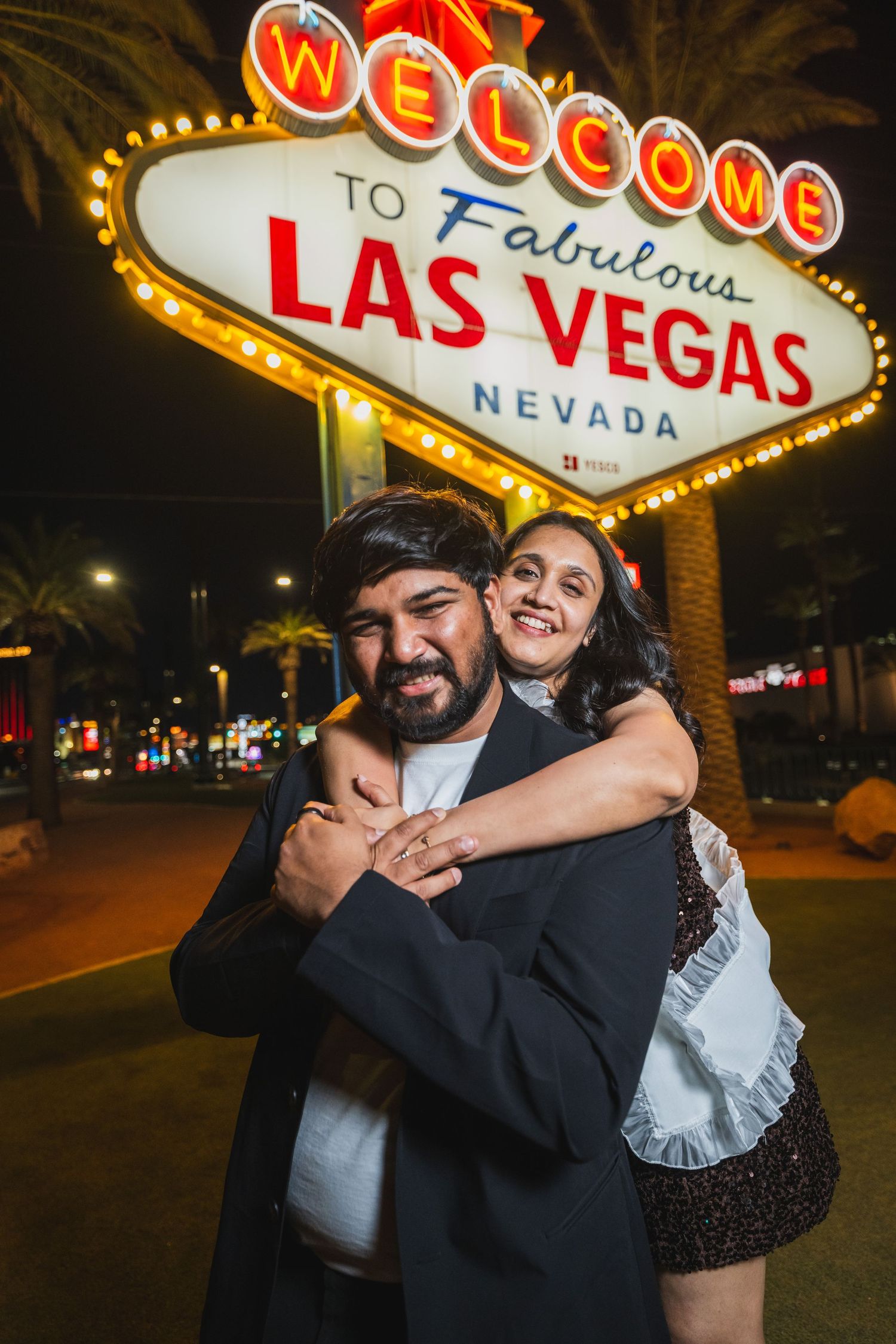 Happy couple embracing in front of the illuminated Welcome to Las Vegas sign at night.