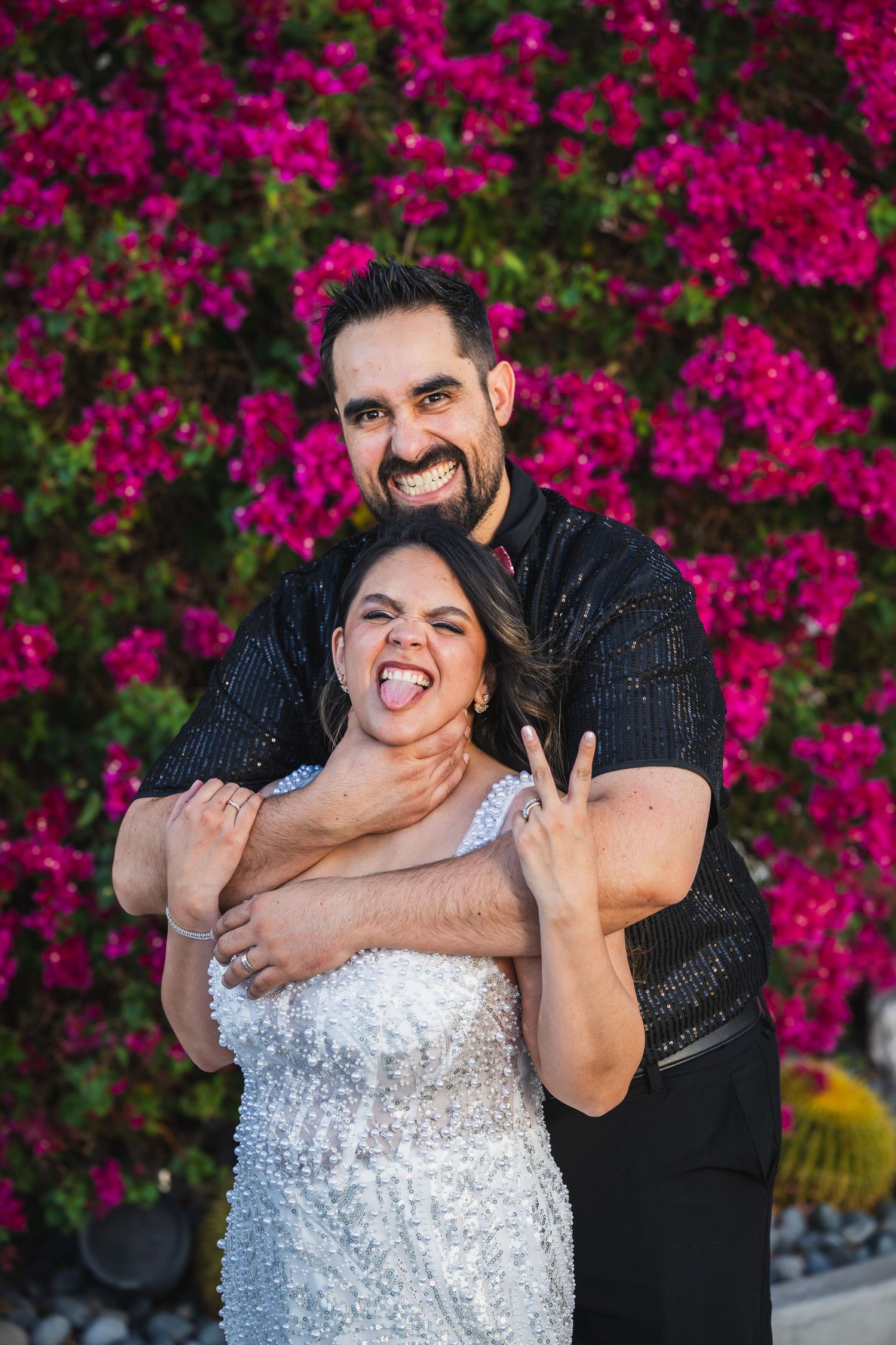 Joyful couple posing playfully in front of vibrant pink bougainvillea flowers outdoors.