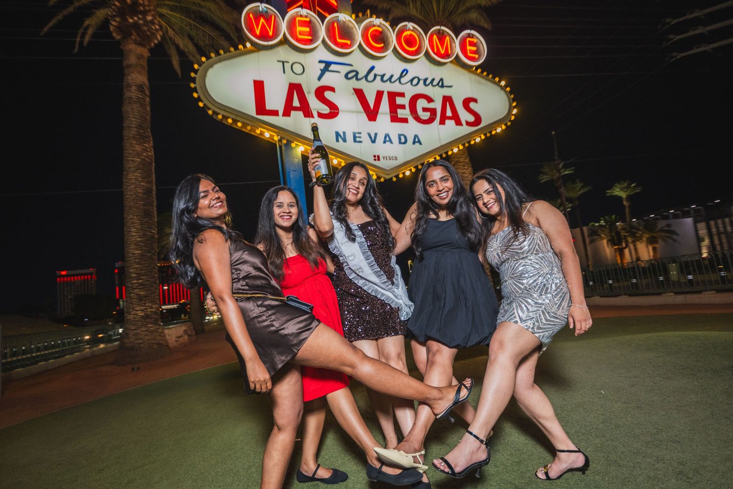 Five women posing and celebrating in front of the Las Vegas welcome sign at night.