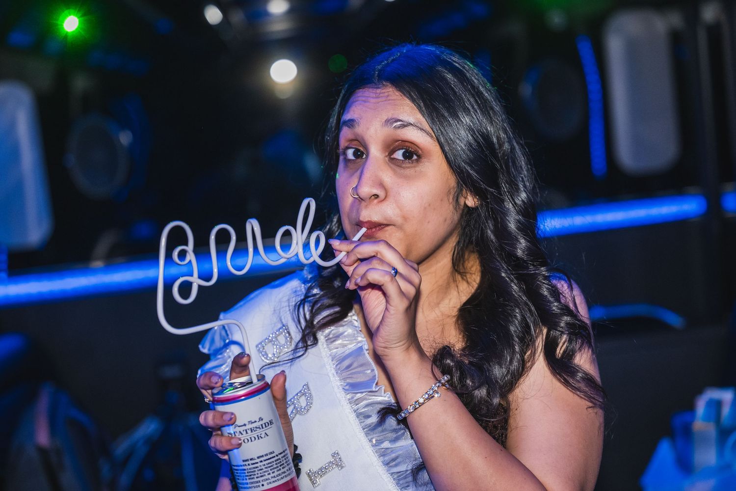 Woman sipping a drink through a straw inside a dark neon-lit party venue.