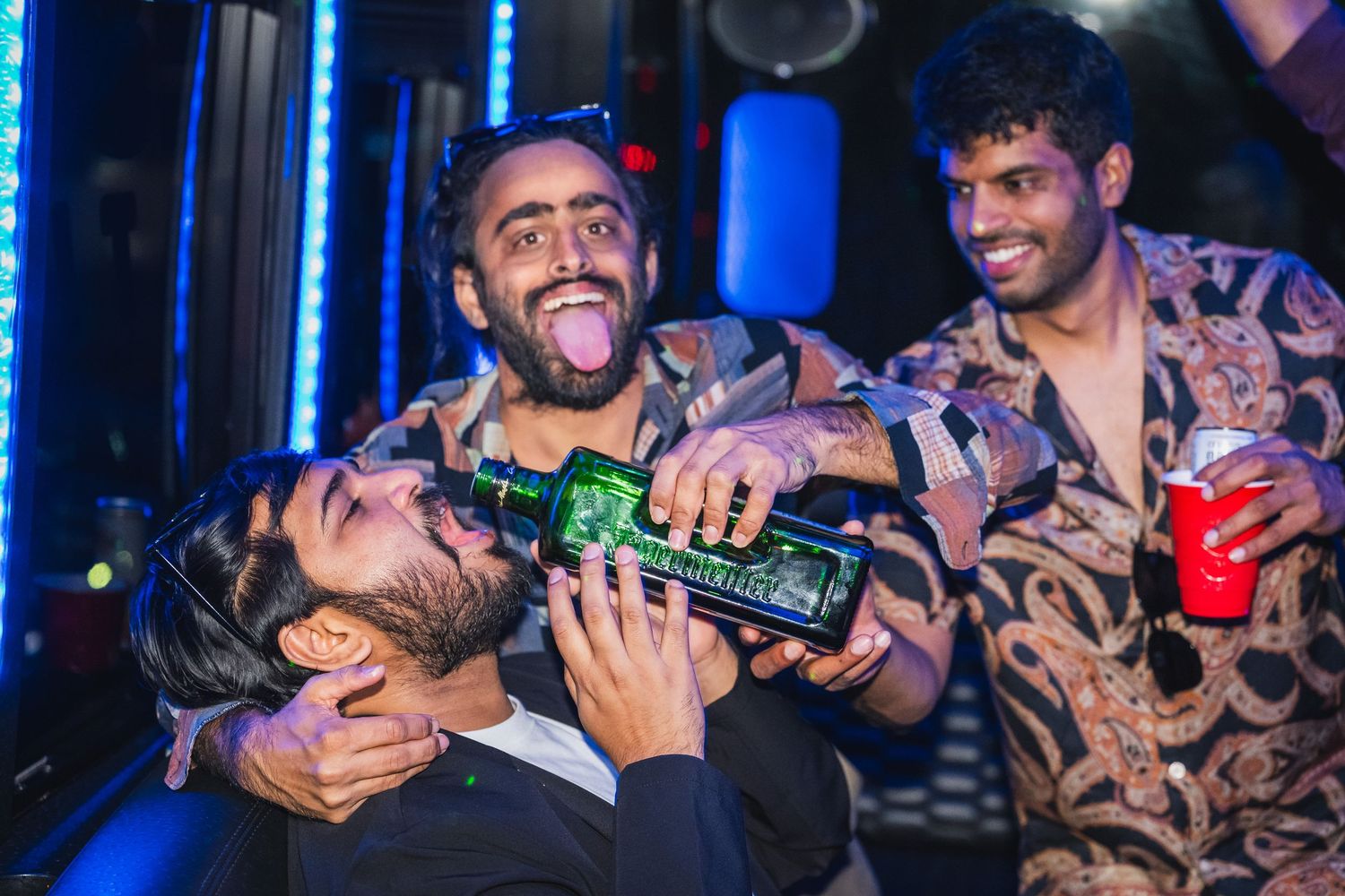 Three men celebrating at a party, one drinking from a green bottle with friends laughing.