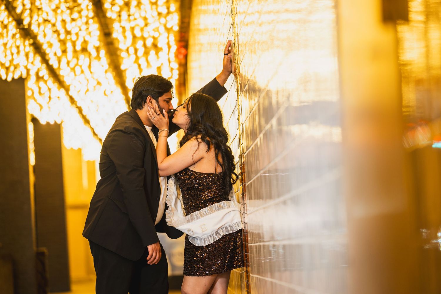 Couple sharing a romantic kiss against a glowing golden wall with string lights in Las Vegas at night.