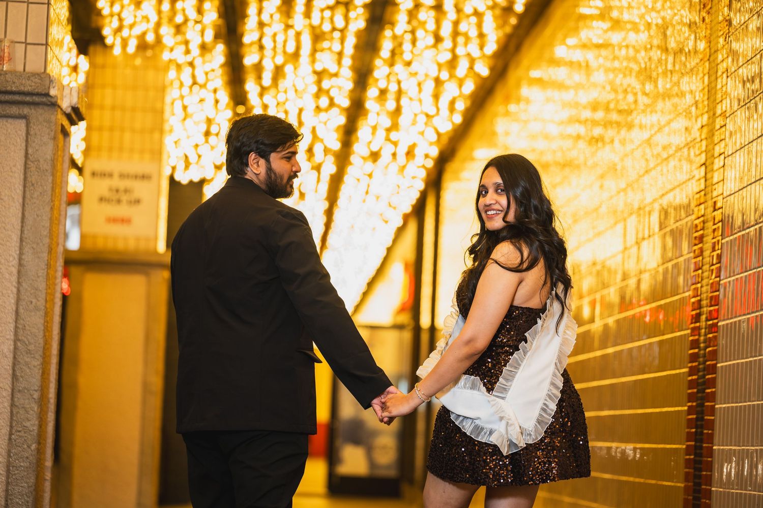 Couple holding hands and smiling under cascading string lights in a warmly lit hallway.