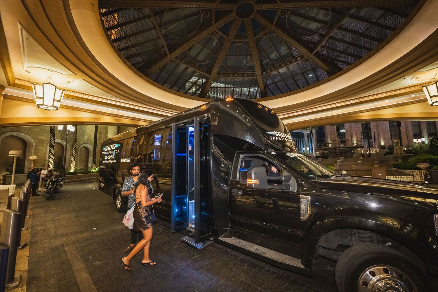 Luxury black party bus parked under ornate hotel entrance with guests arriving at night.