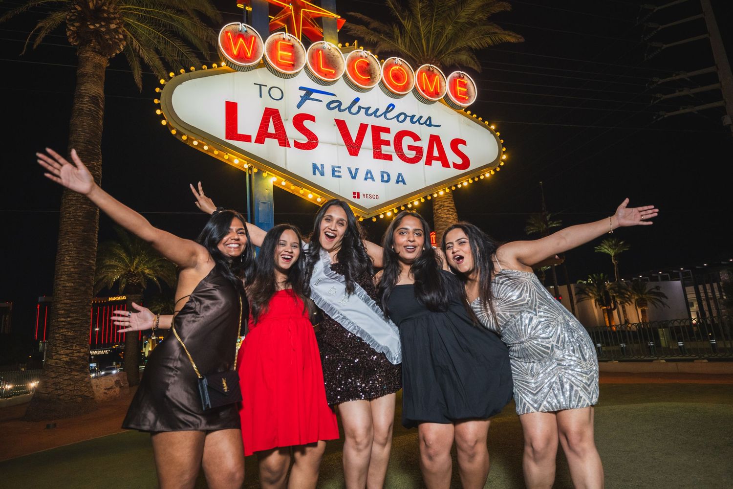 Six women celebrating with arms raised in front of the Las Vegas welcome sign at night.