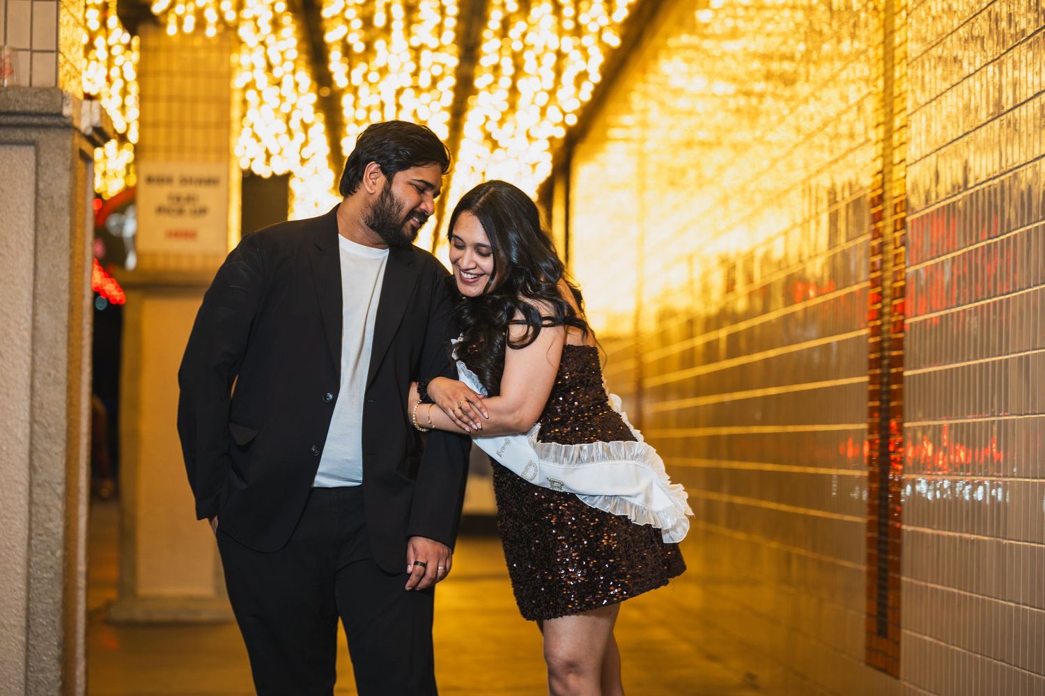 Romantic couple walking hand in hand under warm string lights in a golden-lit corridor.