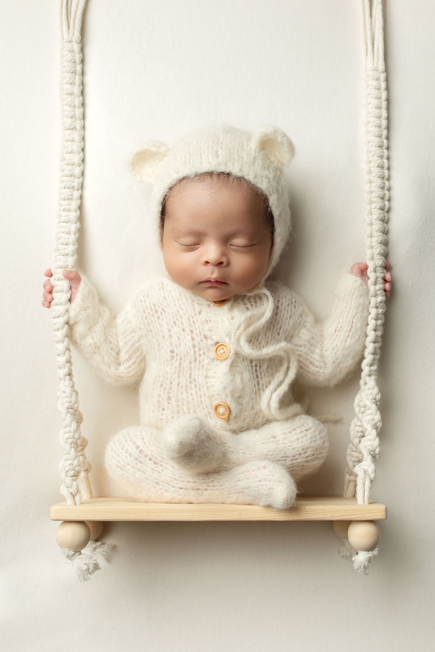 Sleeping baby wearing a cozy white knit outfit with bear ears sits peacefully on a decorative wooden swing.