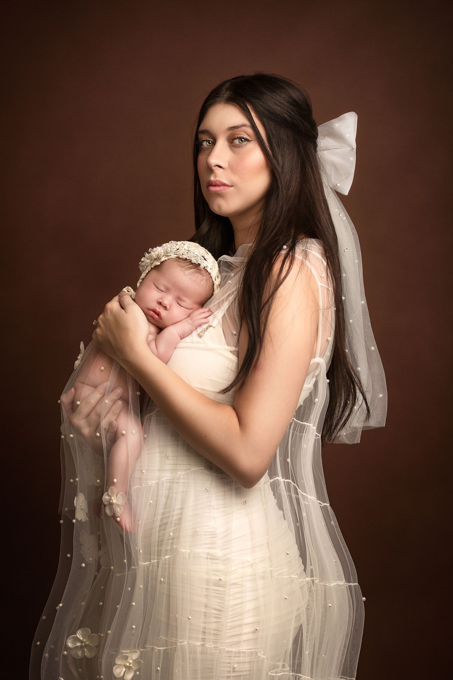 Woman with long dark hair in a cream gown and pearl-beaded veil cradles a sleeping newborn in a floral headband against a brown backdrop.
