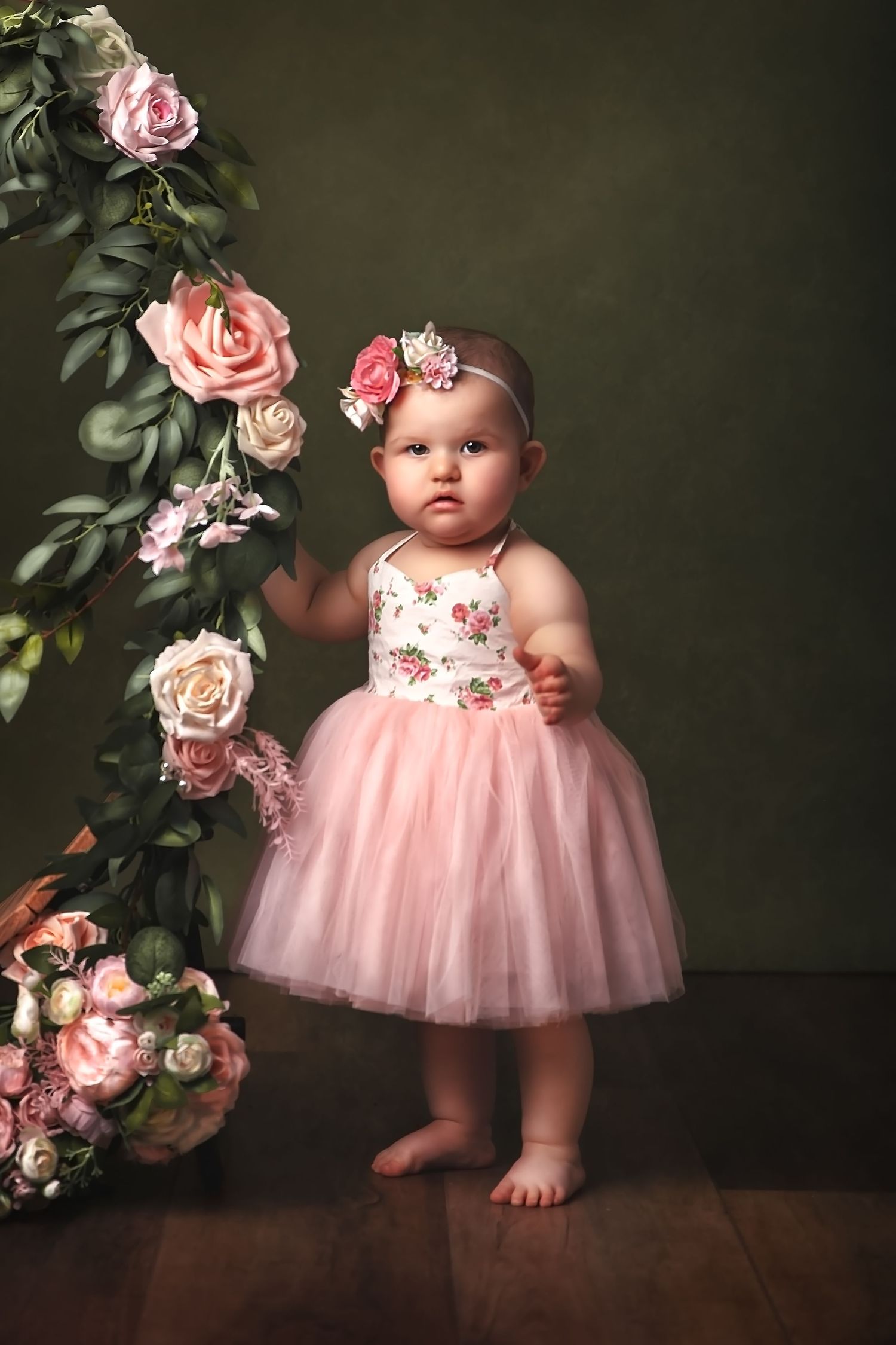 A baby in a pink tulle dress stands next to decorative pink roses against a dark green background in a studio setting.