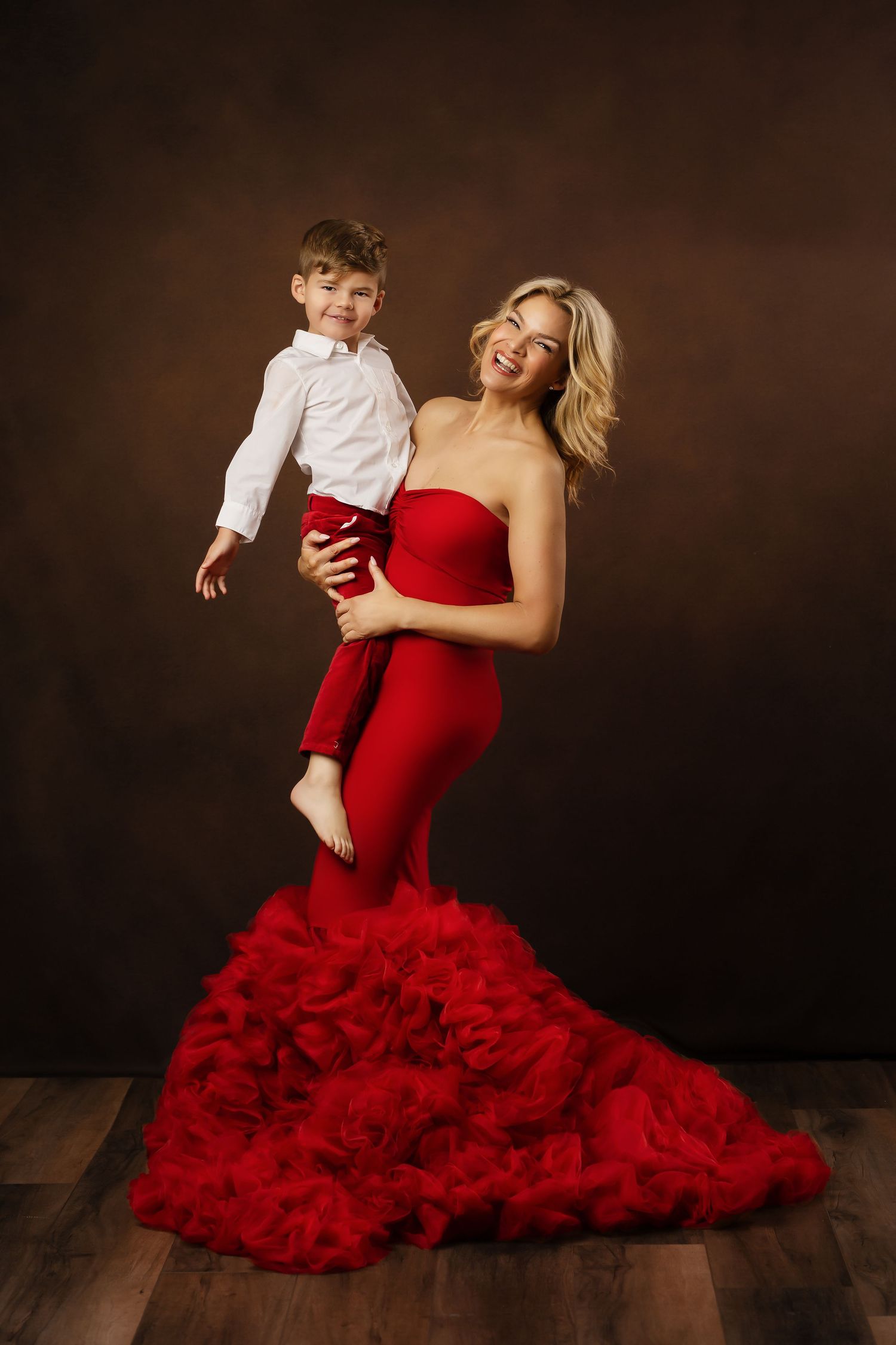 Formal studio portrait showing a person in a stunning red mermaid gown holding a young child in white and red attire.