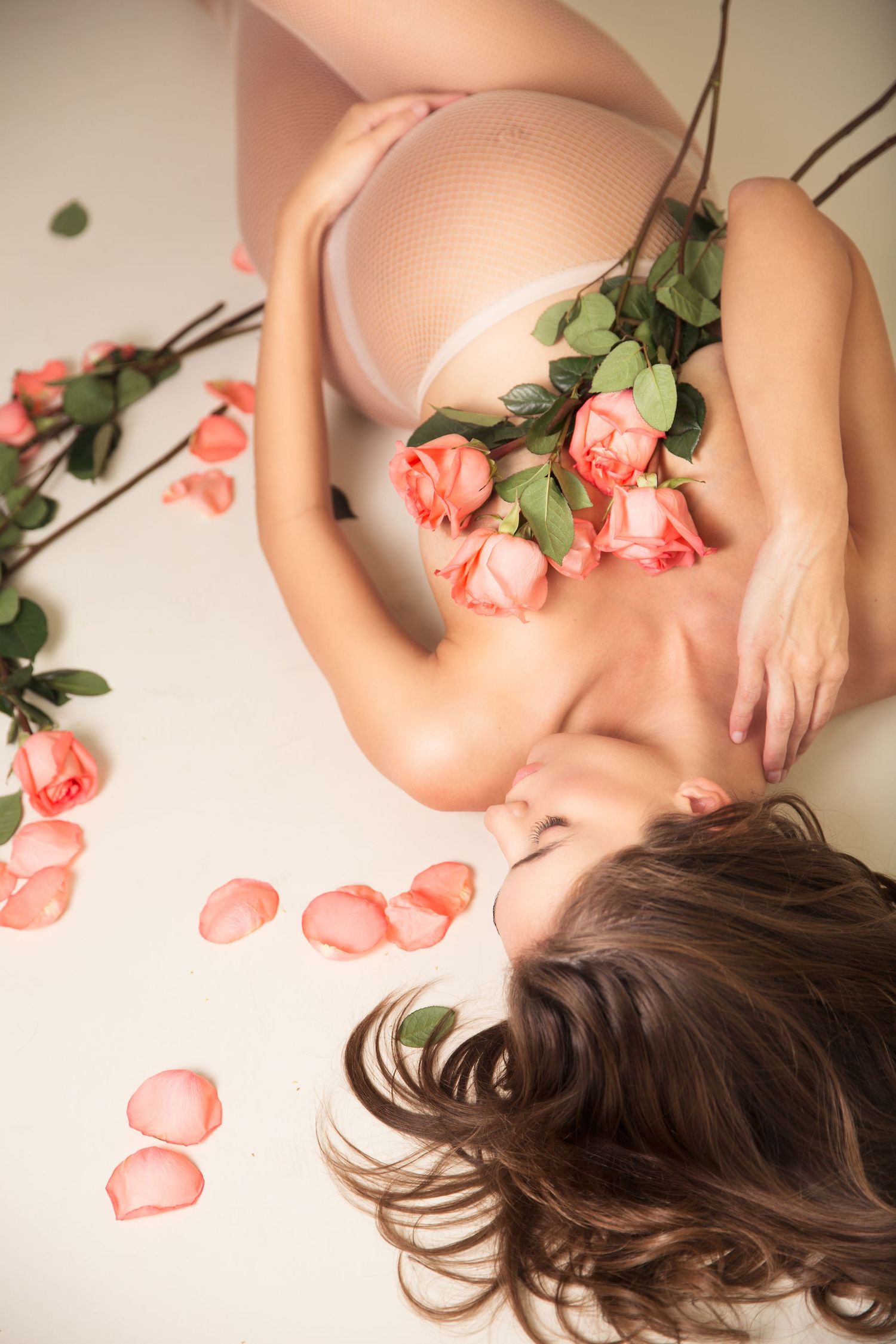 Artistic boudoir photography with soft pink roses scattered on nude-colored fabric creating a romantic, dreamy atmosphere.