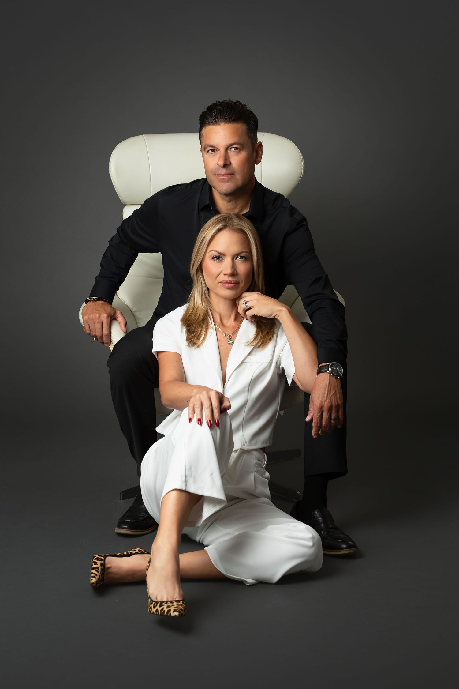 Professional studio portrait of a man in black seated on a white chair beside a woman in a white suit and leopard print flats on the floor.