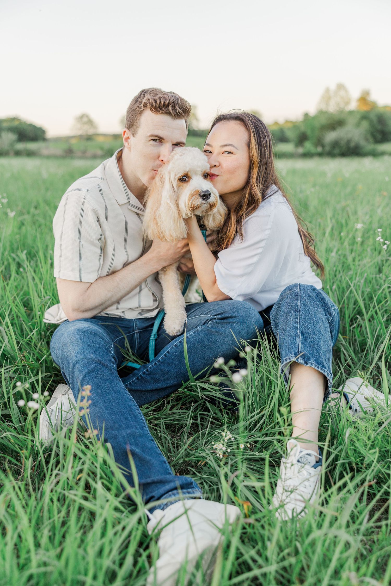 Young couple sitting in green grass field kissing their fluffy golden dog on a sunny day.