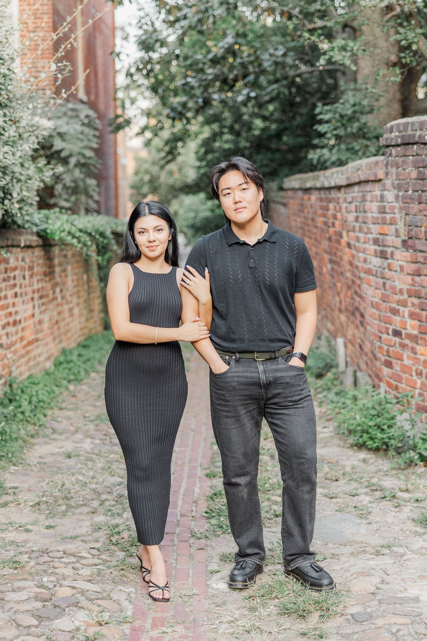 Couple walking and laughing together on a cobblestone path beside a brick wall.