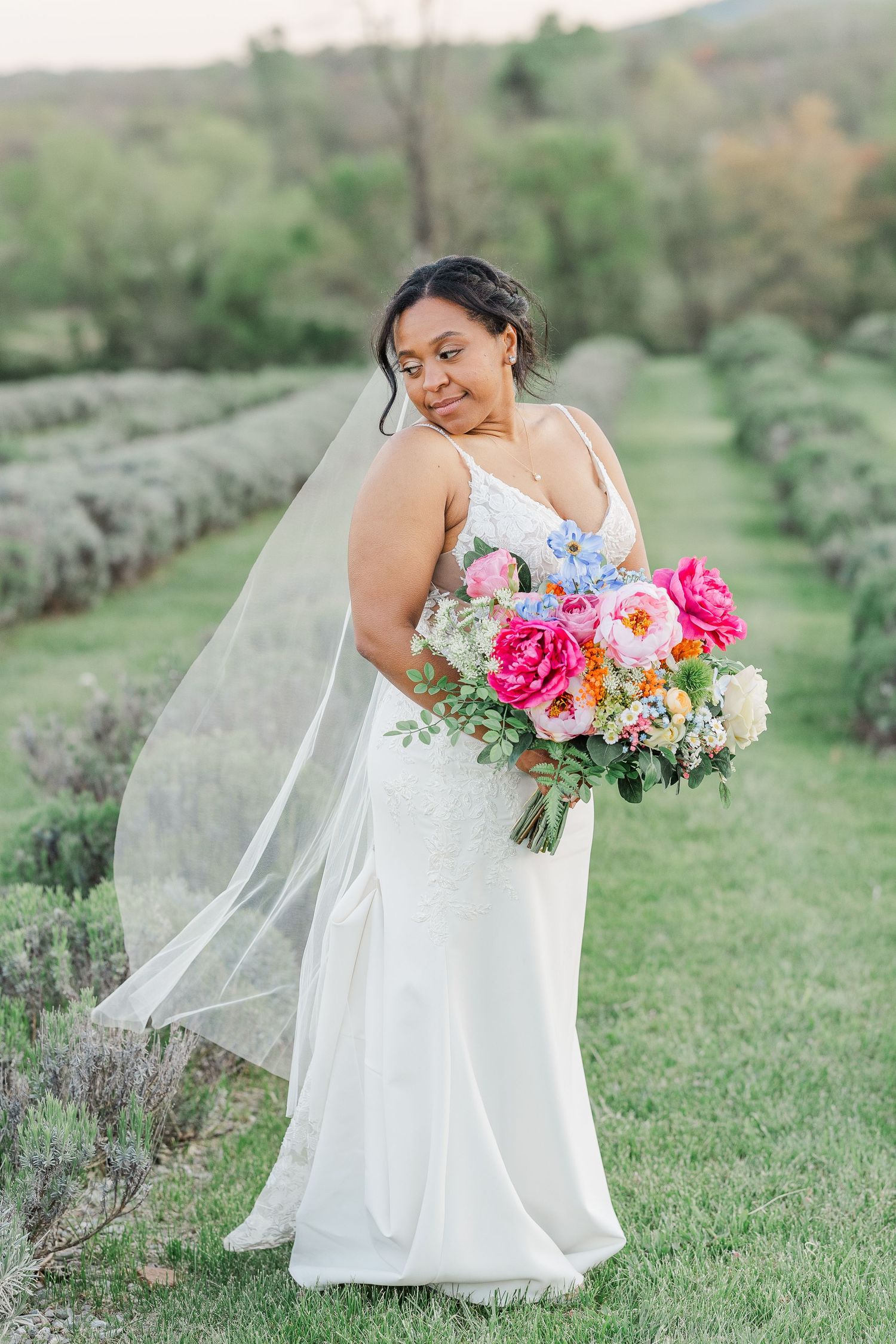 Bride in white dress holds vibrant pink and white floral bouquet in outdoor garden setting.