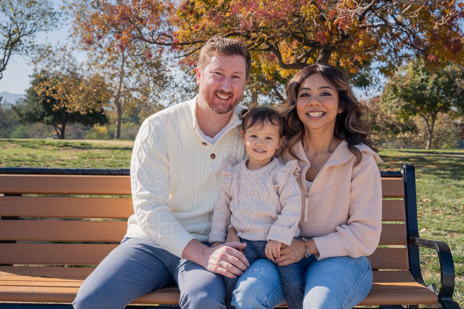 A family wearing coordinated cream and denim outfits sits together on a wooden bench in an autumn park setting.