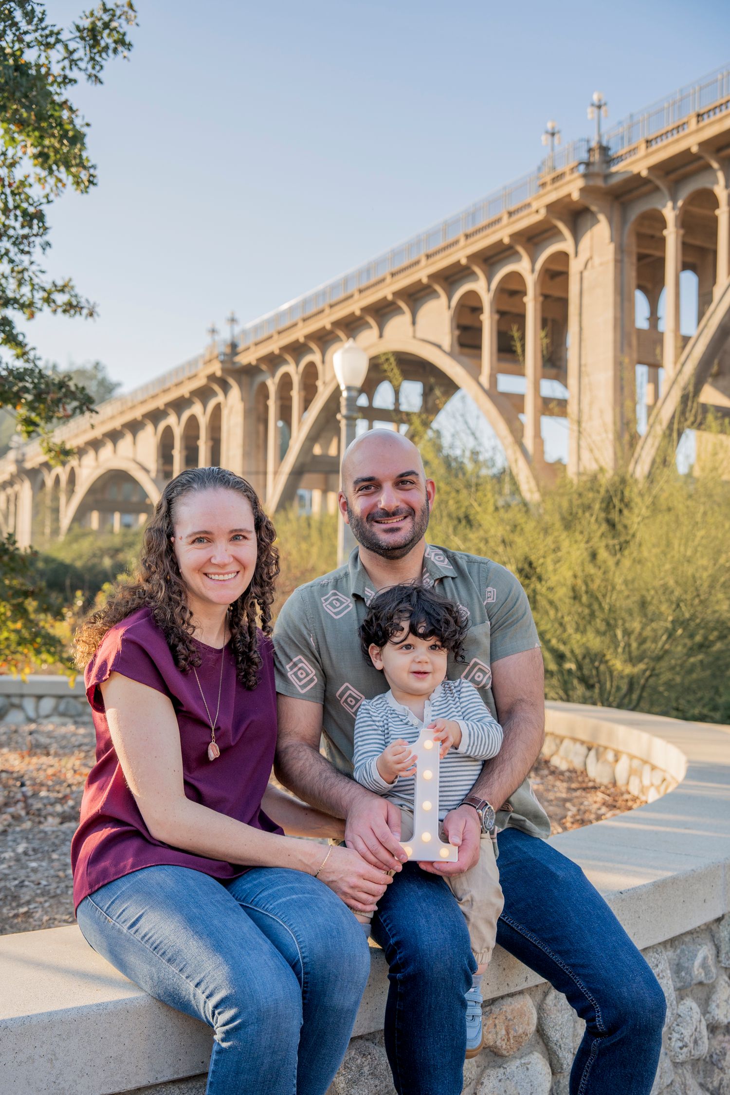 A family sits together on concrete steps with a large arched bridge visible in the background.