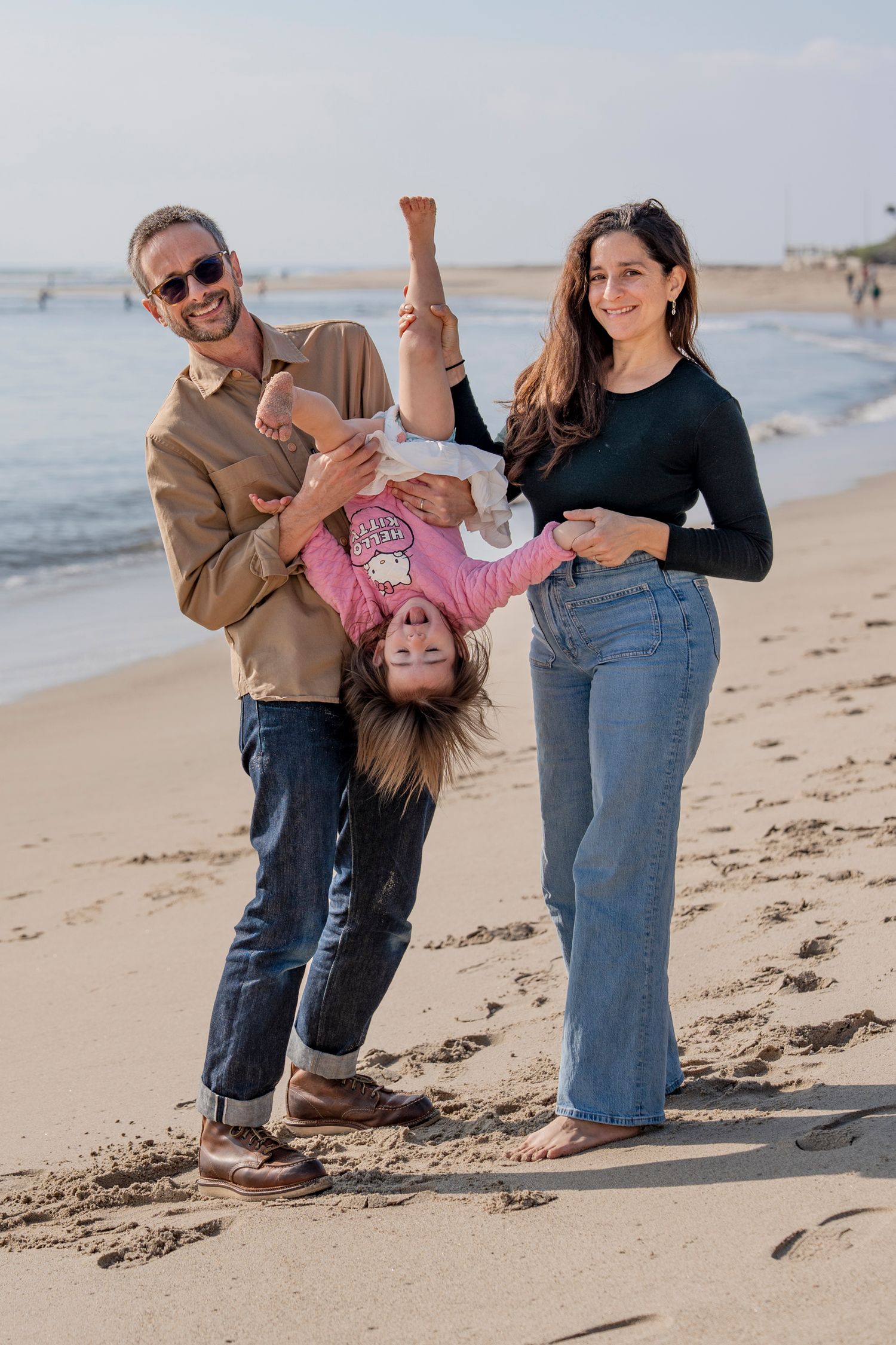 A family plays together on a sunny beach, swinging and laughing while enjoying a carefree moment by the ocean.