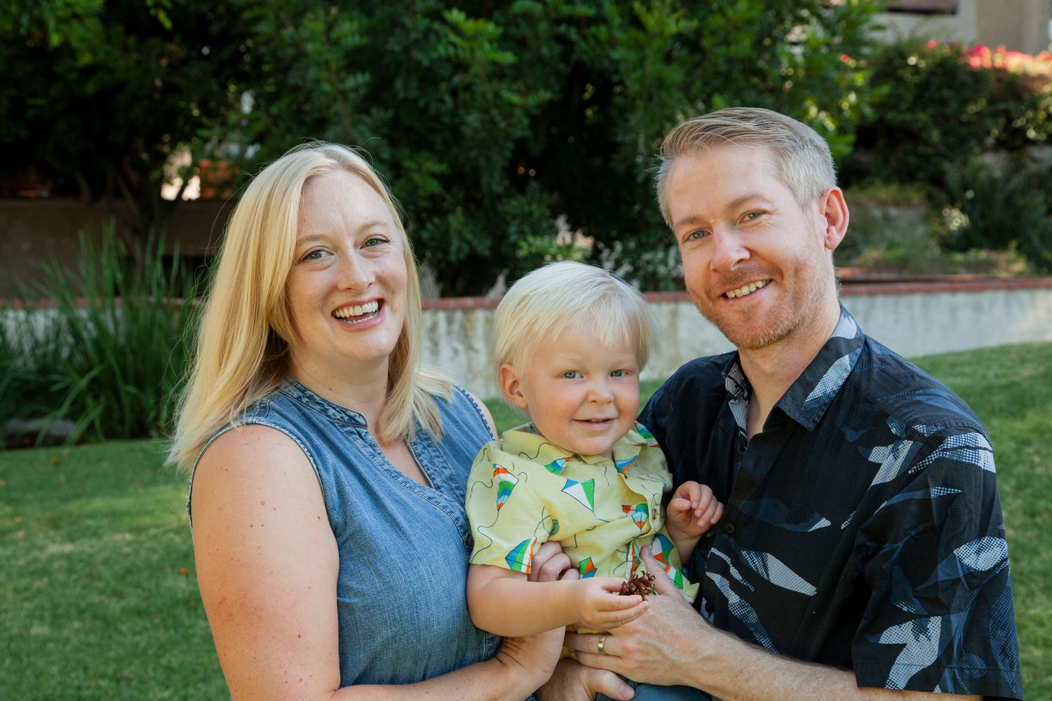 A happy family of three smiles together while sitting on grass in a garden setting.