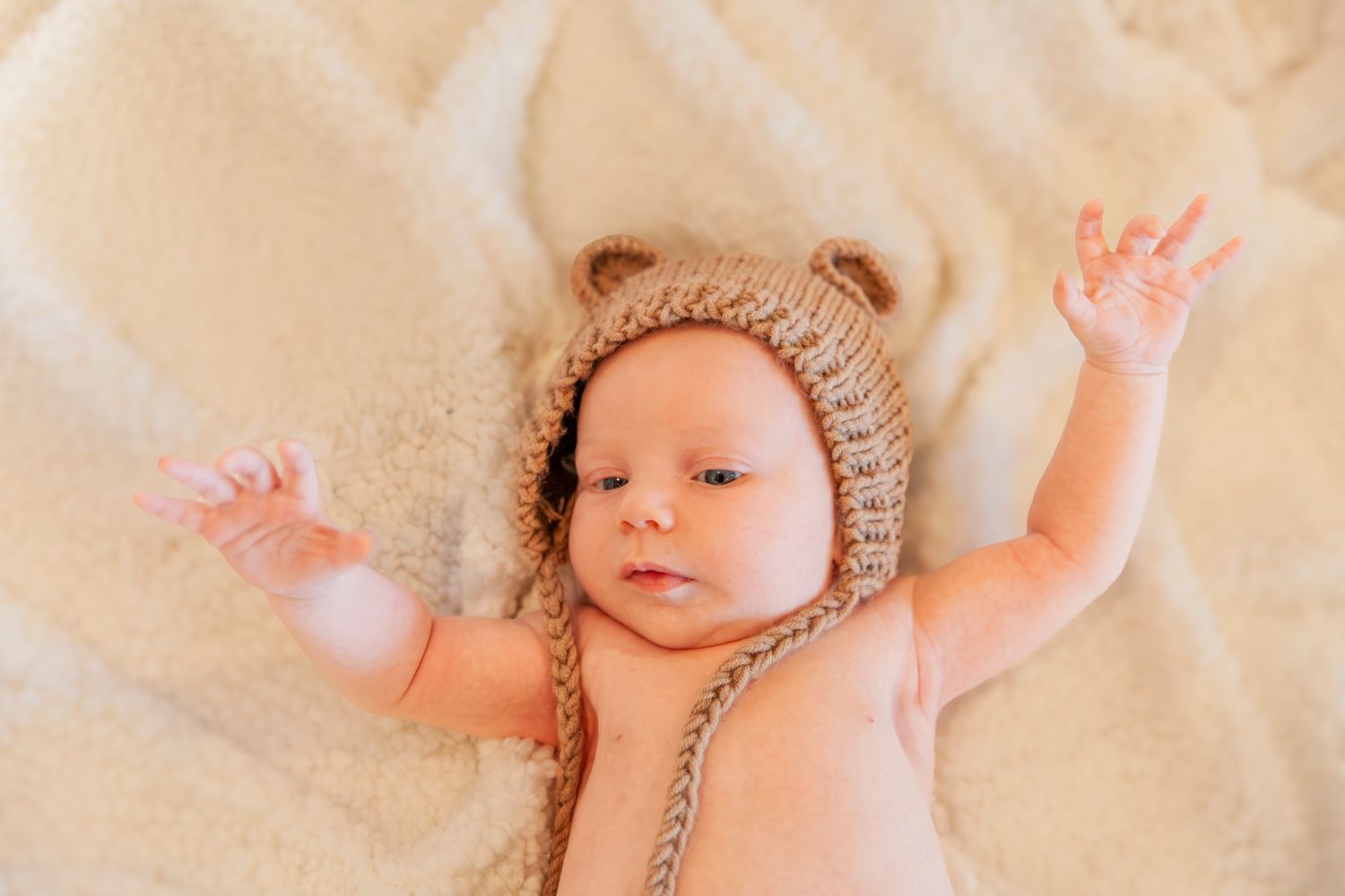 Baby wearing a knitted brown bear hat lies on beige blanket with arms raised.