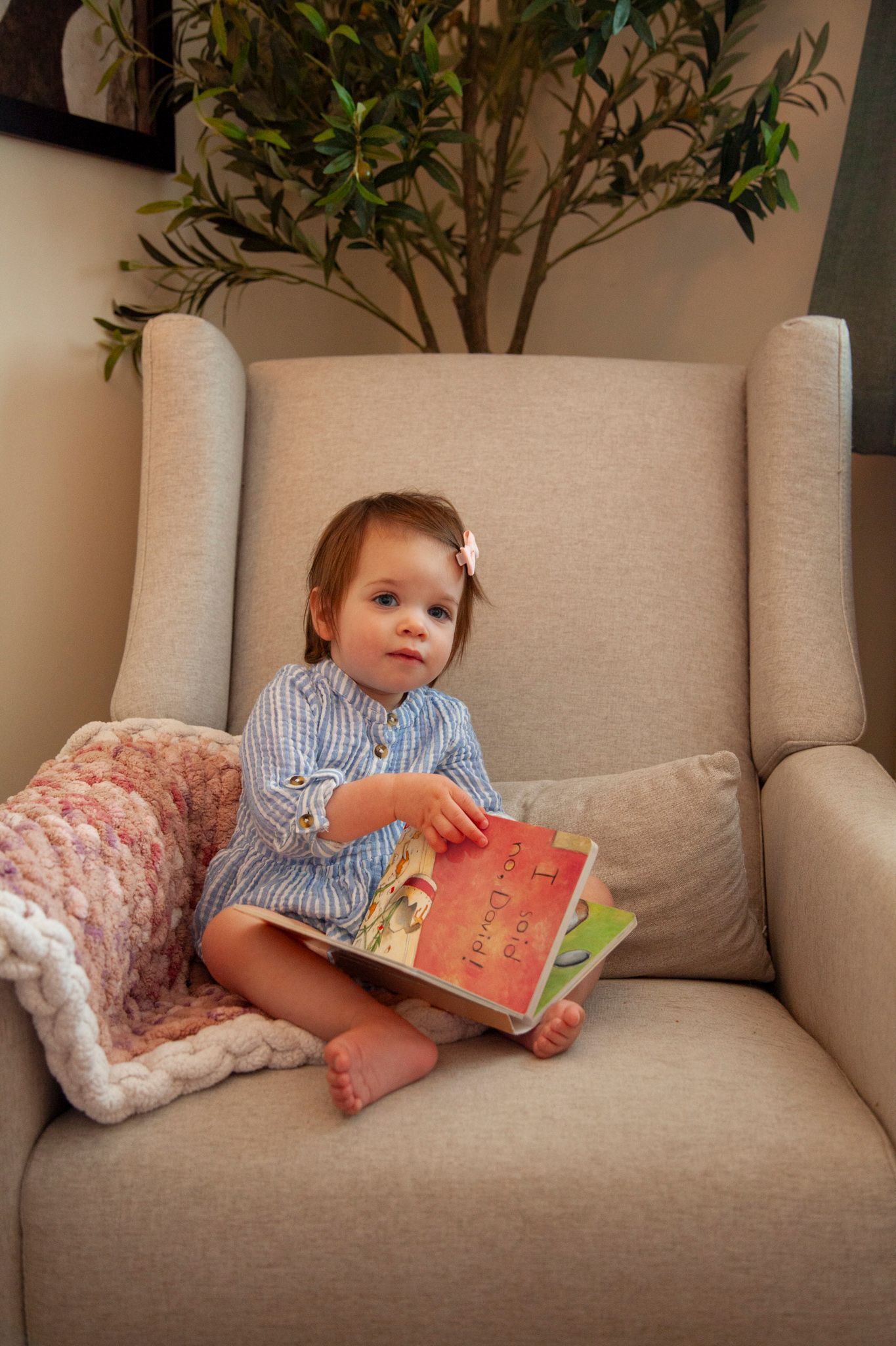 A cozy reading nook features a beige armchair with a soft pink blanket and potted plant in the background.