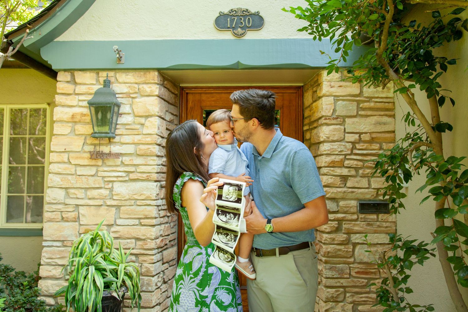 A couple shares a tender moment in front of a stone wall exterior with lush green plants surrounding them.