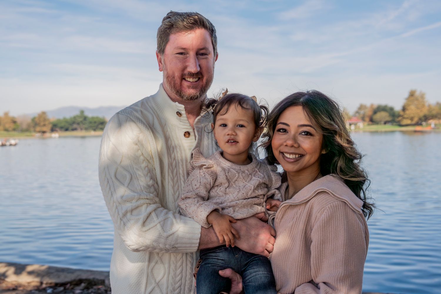 A family enjoys a tender moment together while posing outdoors by a scenic lake on a sunny day.